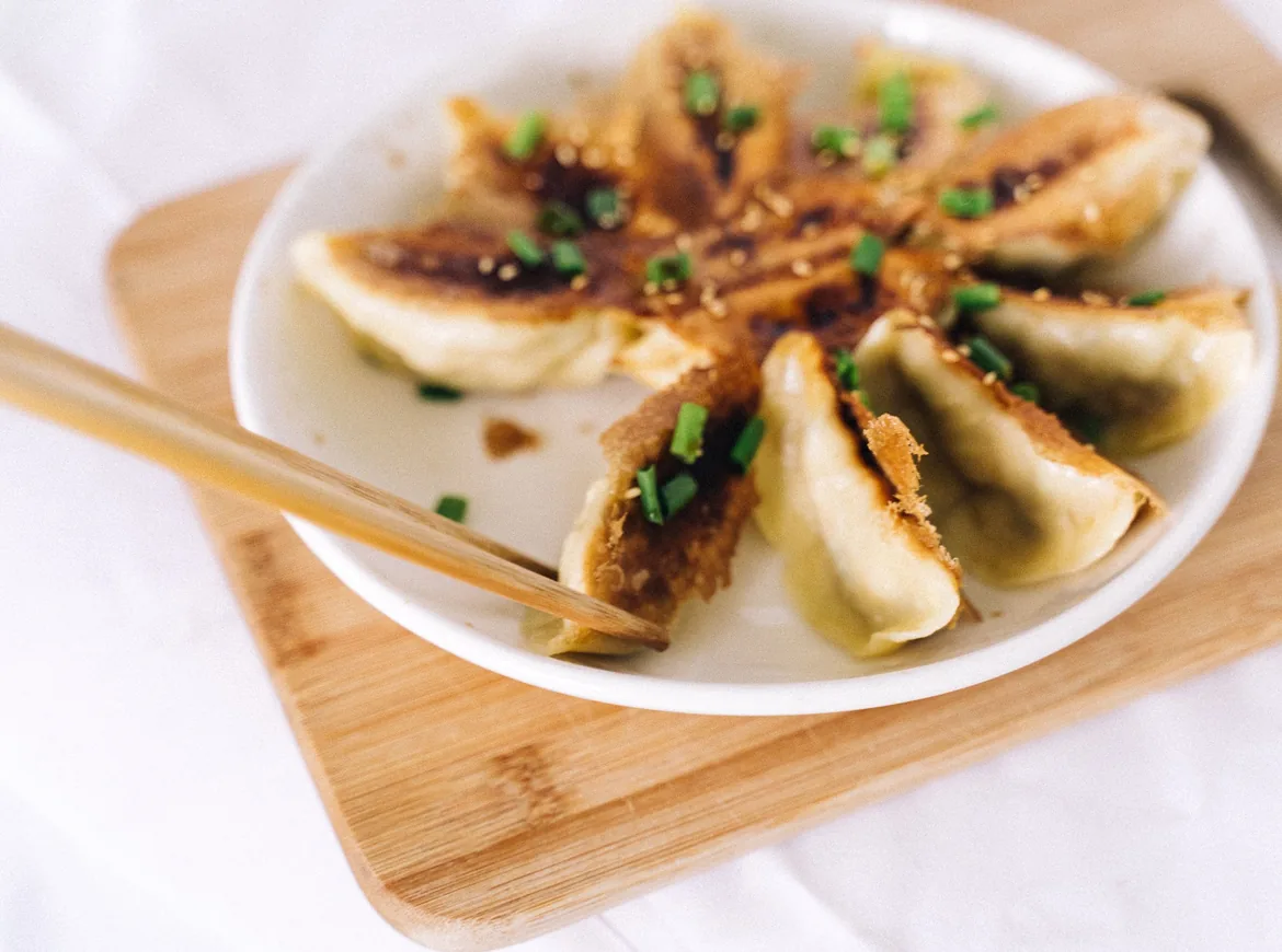 Pan-fried gyoza fanned out in a circle on a white plate set on a wooden board, with chopsticks and a scatter of spring onion