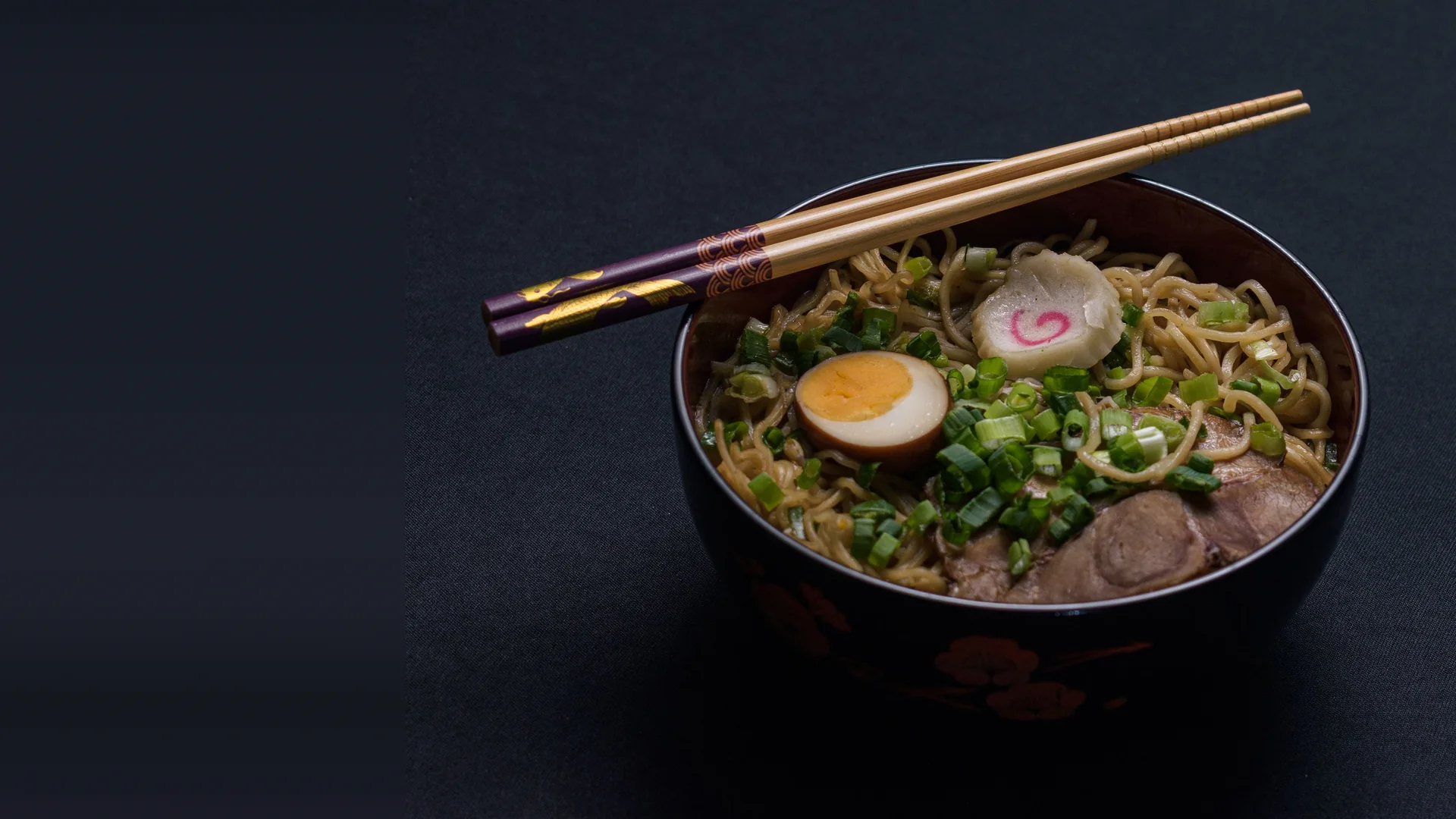A black lacquer bowl of ramen with a halved soft-boiled egg, a pink-swirled fish cake, chopped spring onion and sliced pork, wooden chopsticks resting across the rim, on a dark cloth