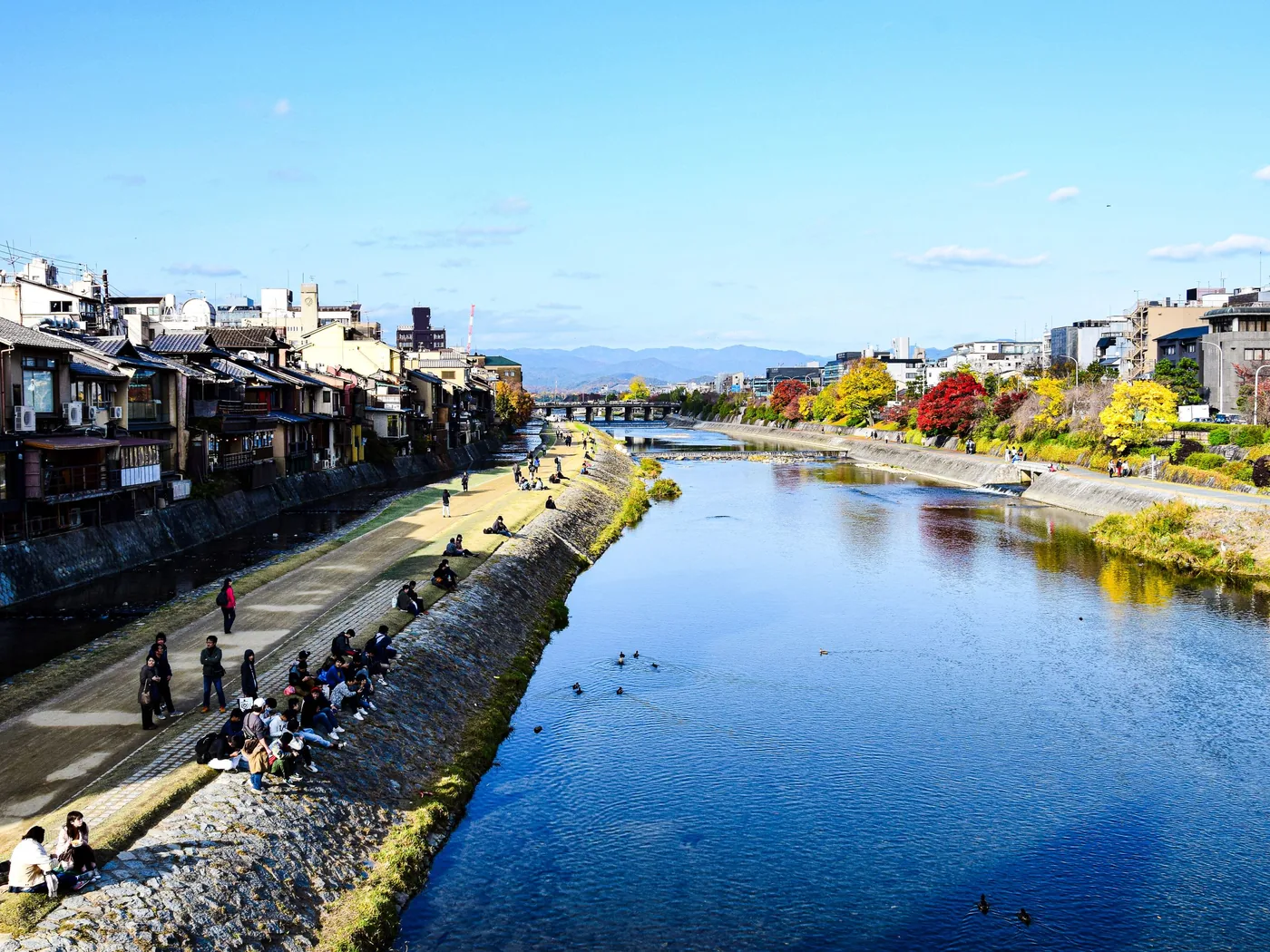 The Kamo River in Kyoto on a sunny autumn day, with wooden riverside buildings on the left bank and people sitting along the stone embankment