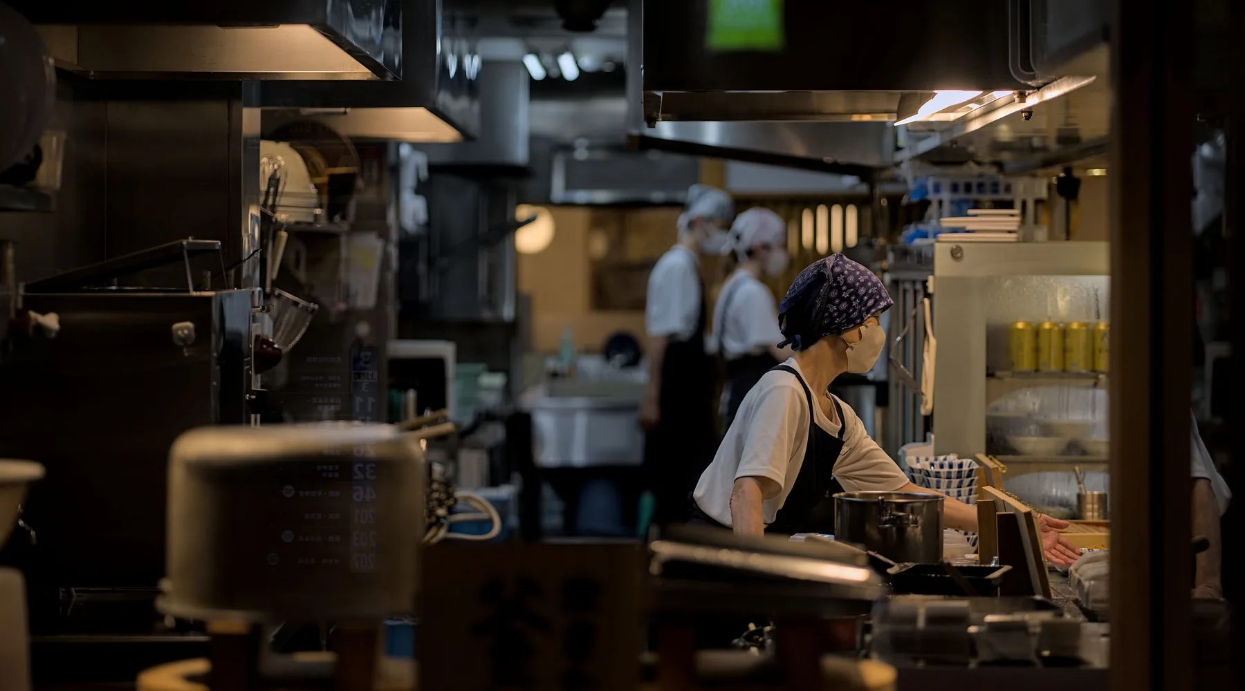 A busy open kitchen in Kyoto at night, cooks in aprons and headscarves working at the counter beneath hanging lights