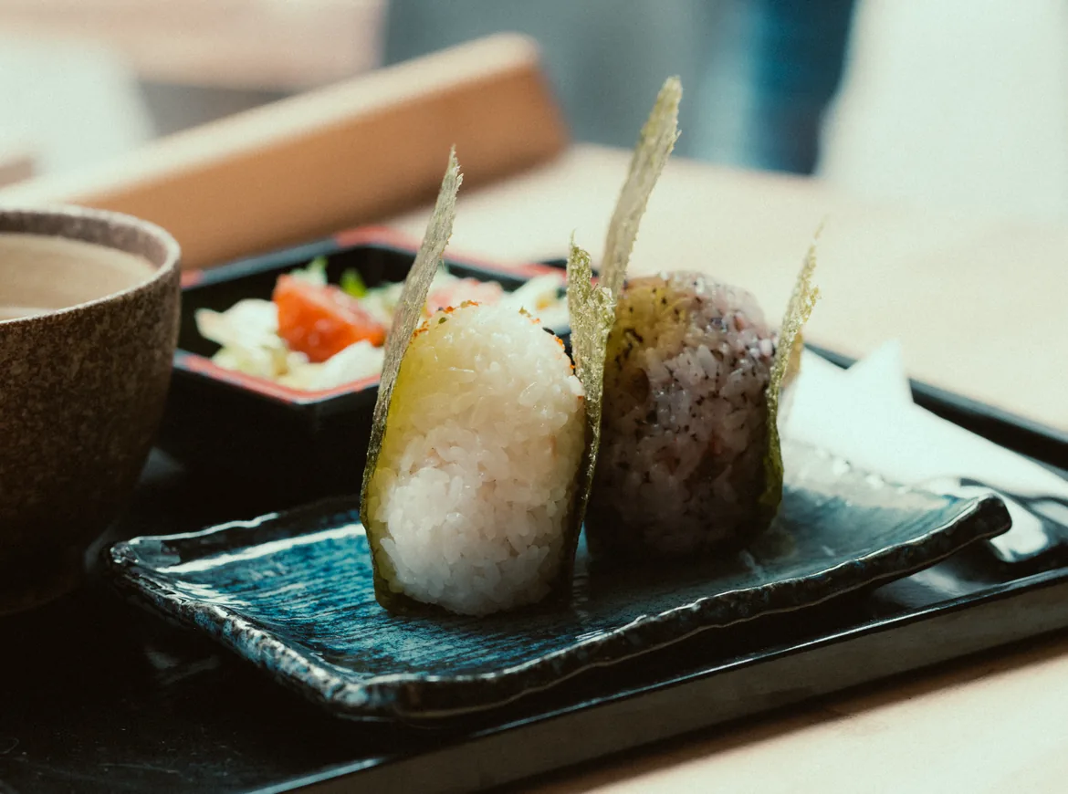Two onigiri wrapped in strips of nori on a blue-glazed plate beside a cup of tea