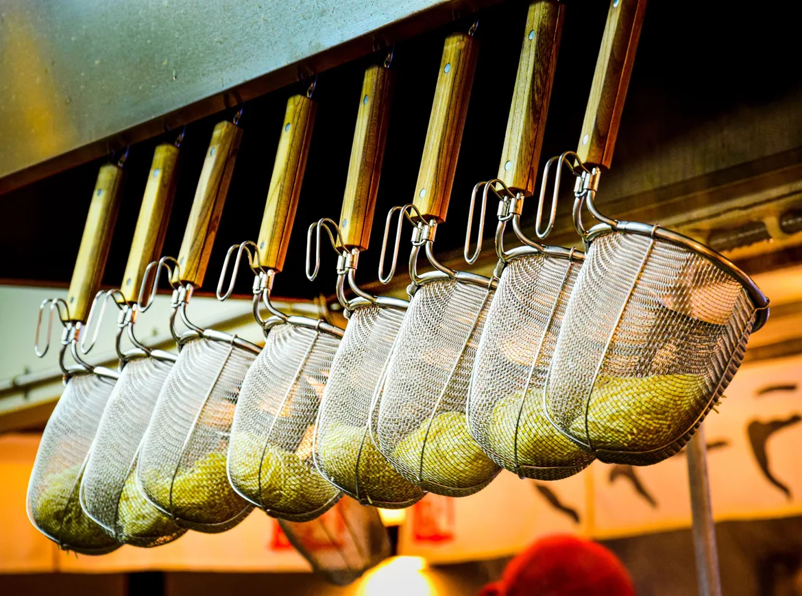 A row of long-handled noodle strainers hanging in a ramen shop kitchen, lit amber from below